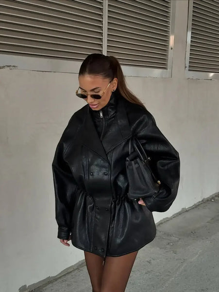 A woman wearing an oversized black faux-leather jacket with a drawstring waist, styled with a black mini dress, sunglasses, and a black handbag, standing against a concrete wall.